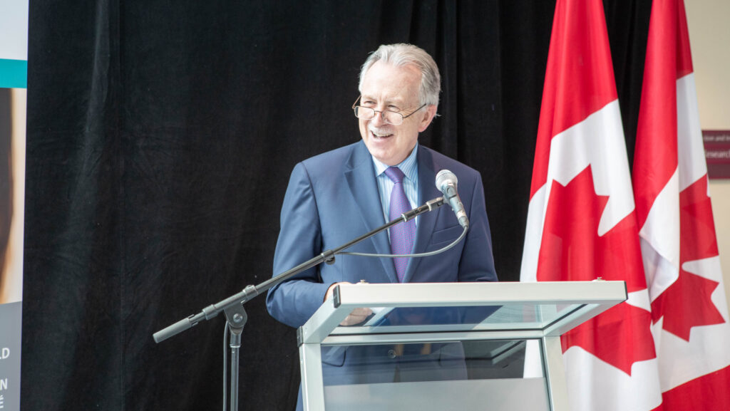 Paul O’Byrne smiles at a podium with a Canadian flag to his left.