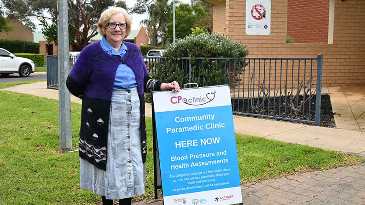 An older adult woman stands beside a sign advertising the CP at clinic program, outside of a community centre.