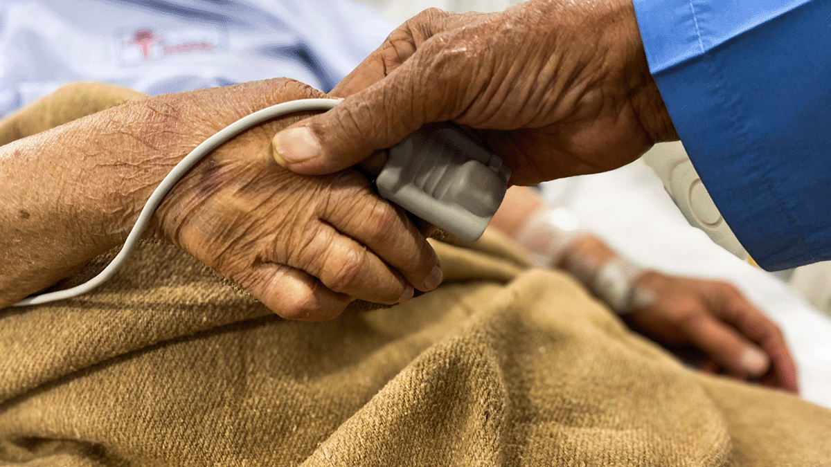 An older couple holds hands. One of them has a heart rate monitor on their finger with a blanket draped over their lap while they sit in a bed.