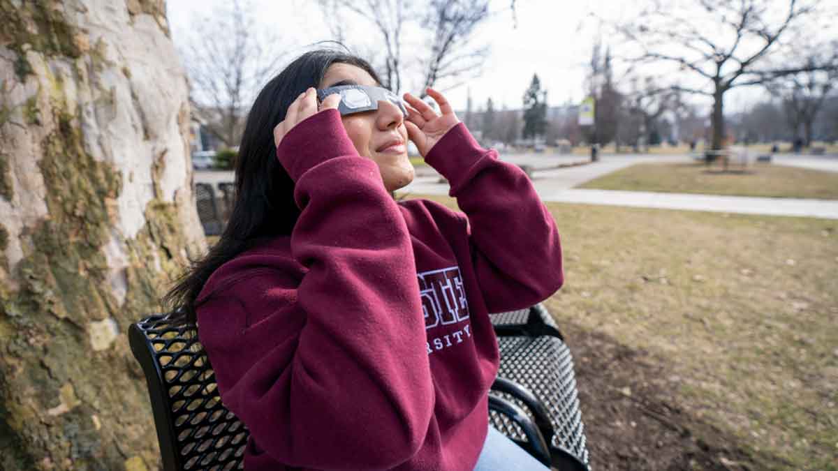 A student in a McMaster sweatshirt wearing solar eclipse glasses and looking up at the sky