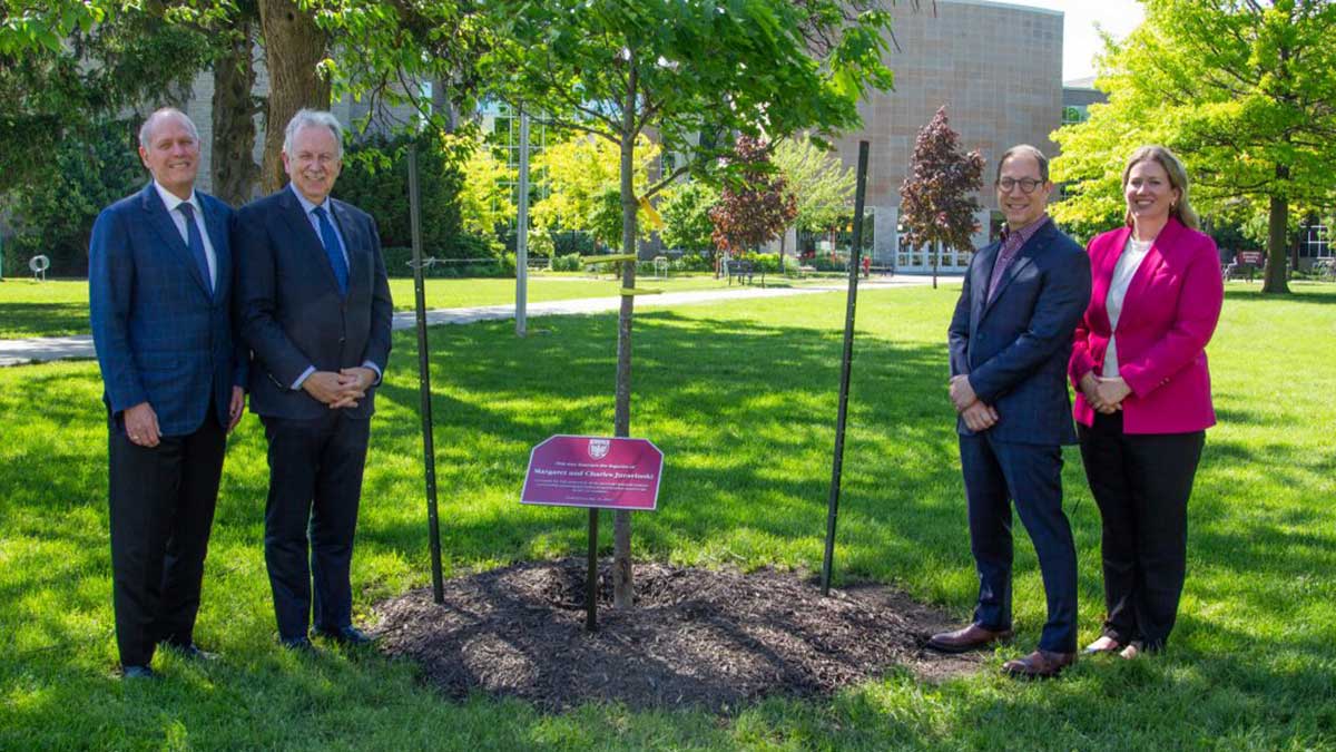 Four individuals, standing near a newly planted red oak tree, commemorate the memory of Hamilton philanthropists Charles and Margaret Juravinski. A commemorative maroon plaque is visible in front of the tree.