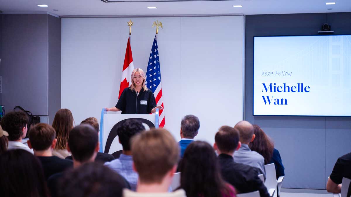 Michelle Wan is wearing a black shirt and is standing at a podium between a Canadian and American flag. She is facilitating a traditional one minute personal pitch to past Cansbridge Fellows.