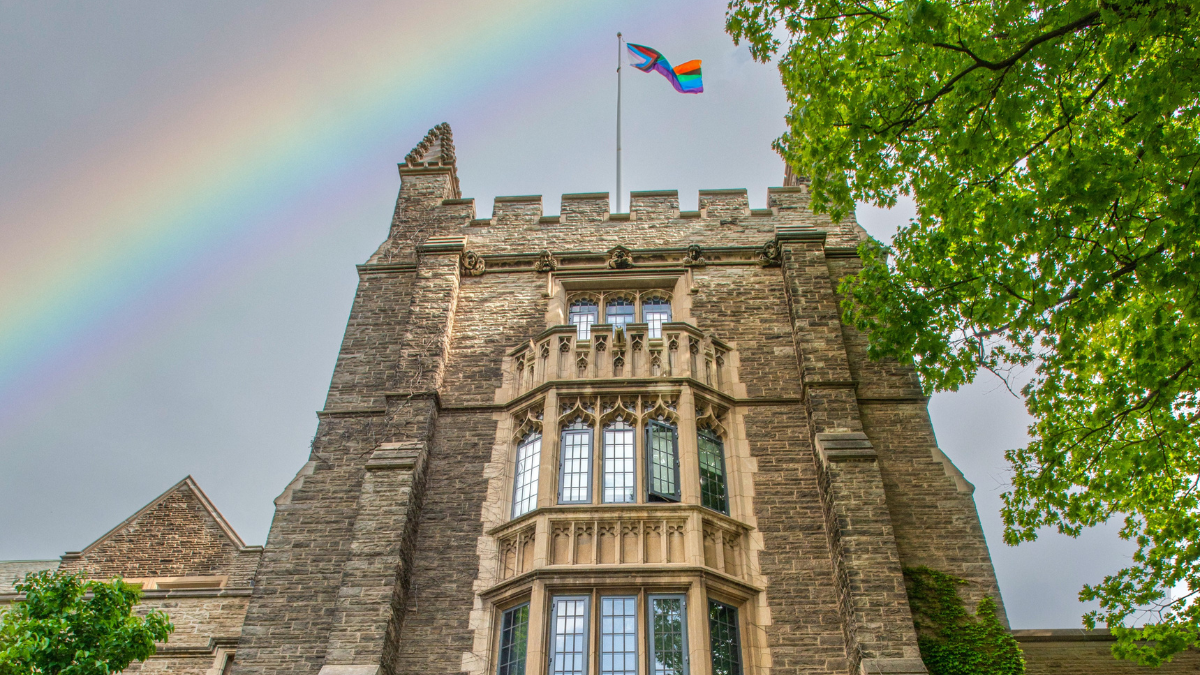 Pride flag raised over University Hall, with a rainbow in the background.