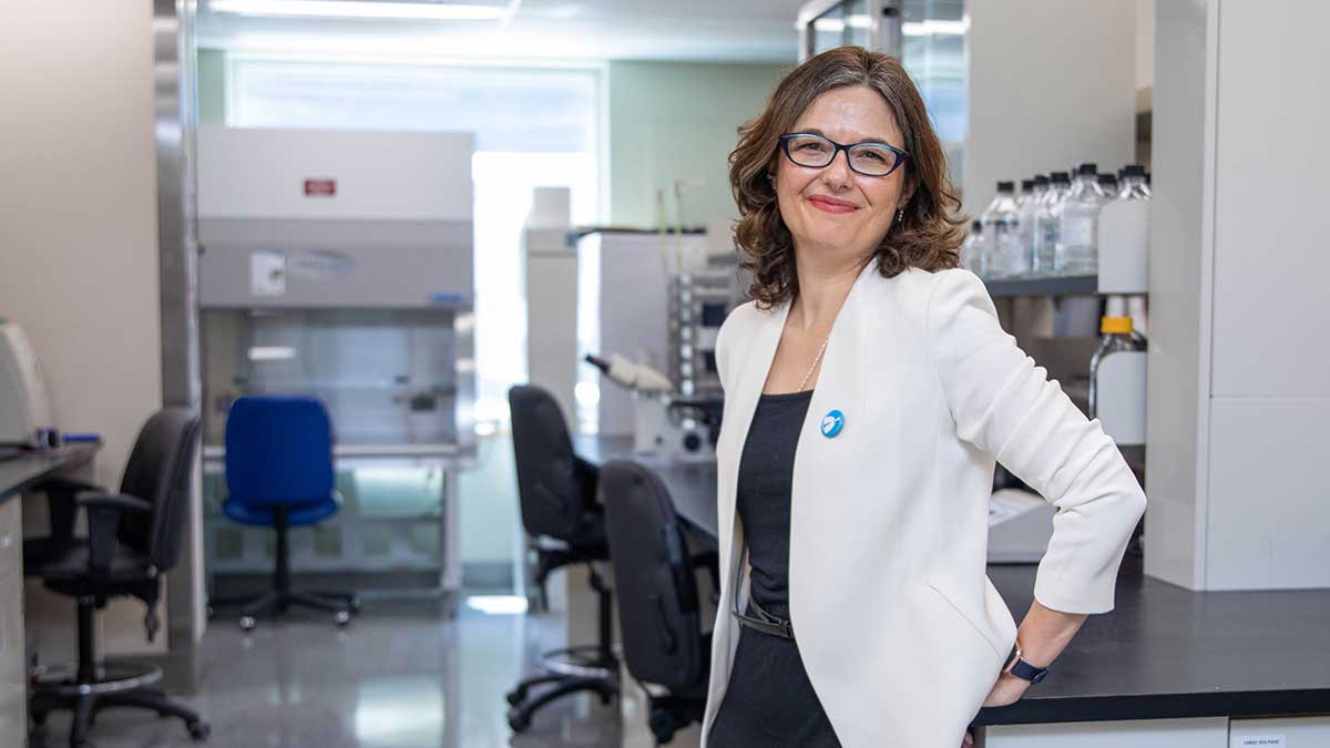 Dawn Bowdish stands in her lab leaning against a lab bench. She is wearing glasses and a white blazer with a blue pin on her left lapel.