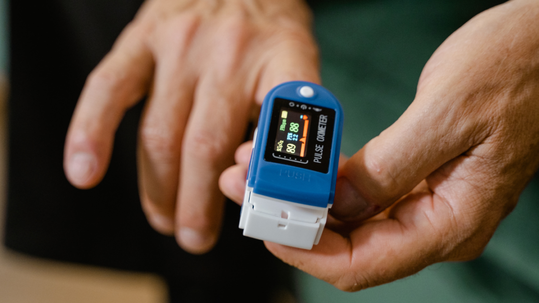 Two hands in frame, the left hand is guiding a blue pulse oximeter onto a finger of the right hand.