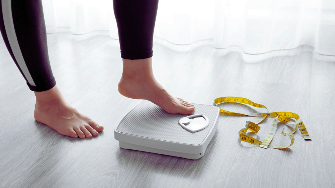 A woman stands barefoot on a scale with a measuring tape sprawled across the floor in the background.