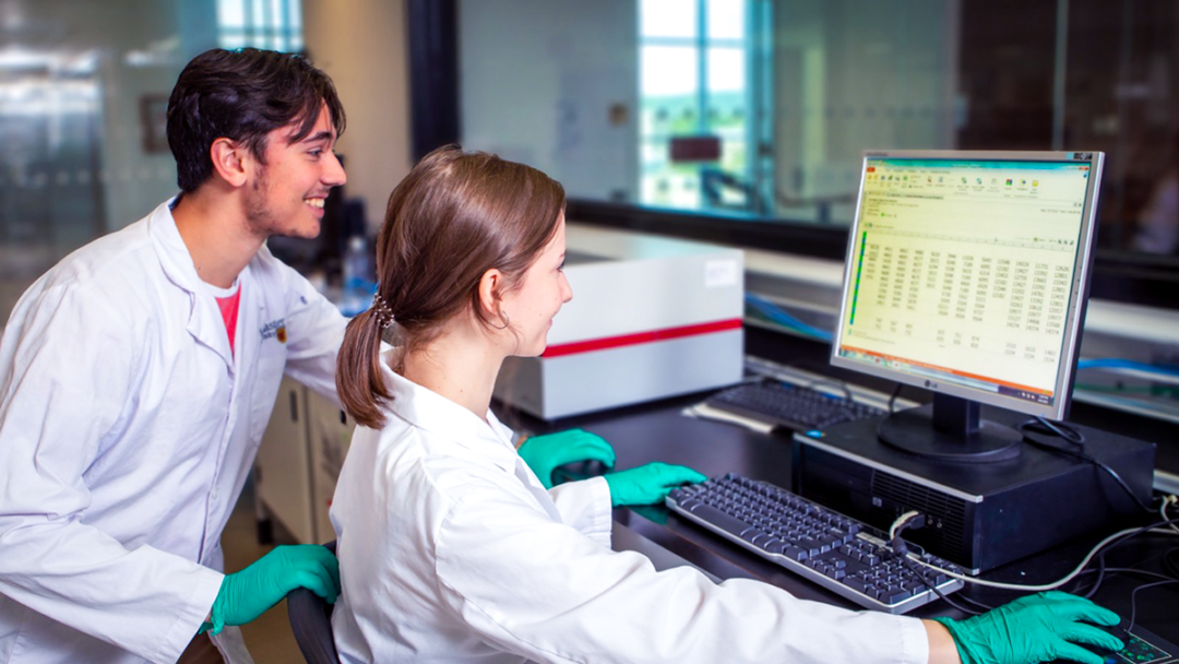 Smiling McMaster University researchers in white lab coats convene around a computer screen to review lab results