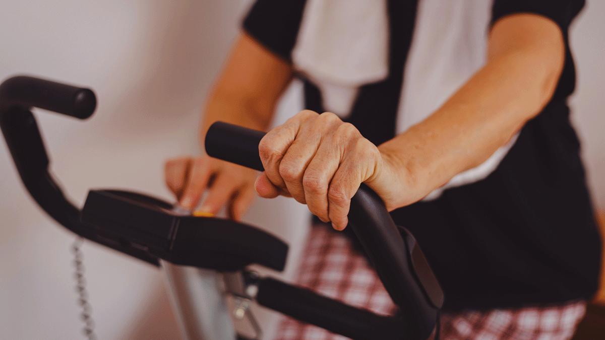 A senior woman can be seen gripping the handle of an exercise bike. A towel is draped around her neck.