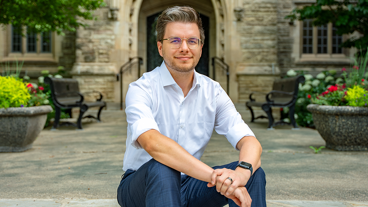 Mike Ayers is wearing a white button up shirty and dark blue slacks. He is sitting on steps with his arms on his knees in front of a building on the McMaster campus.