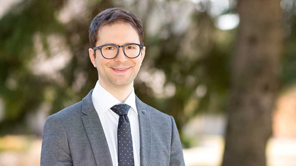 A photo of Matthew Sibbald smiling to camera. He is wearing a grey suit, black tie with polka dots and glasses. He is standing with foliage in the background.