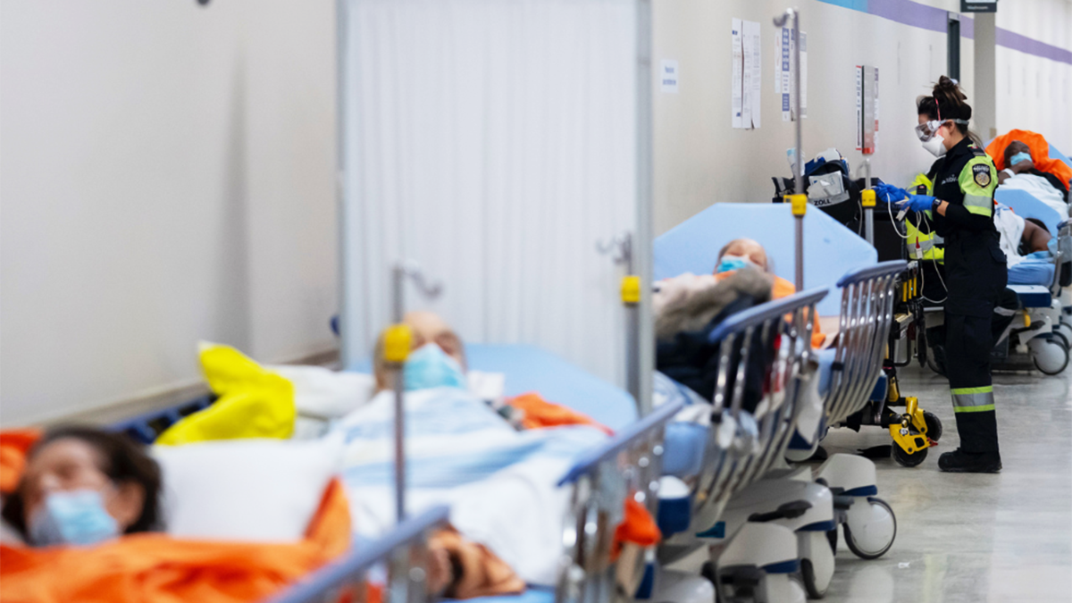 A first responder stands at the side of a patient on a hospital gurney, in a line of such patients on gurneys.