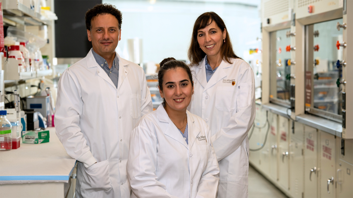 Two researchers stand on either side of a third seated researcher, all smiling and wearing lab coats, inside a lab.