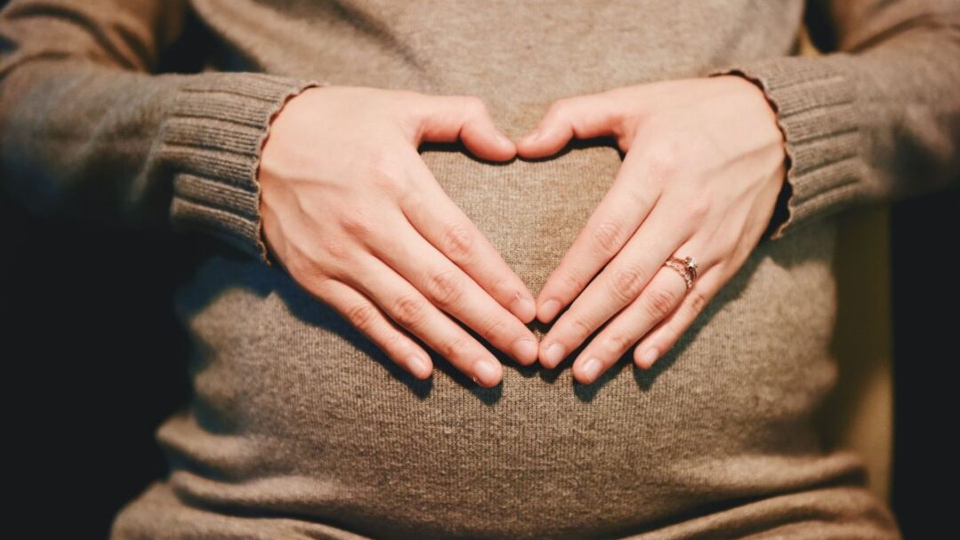 A person wearing a brown top is holding their hands in the shape of a heart on top of their pregnant belly.