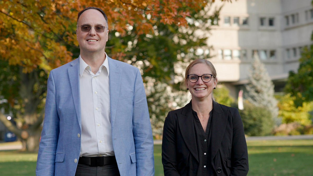 Ray Truant is on the left standing in front of fall foliage and wearing sunglasses and a blue blazer. Tamara Maiuri is on the right in a black shirt and blazer, in front of a building.