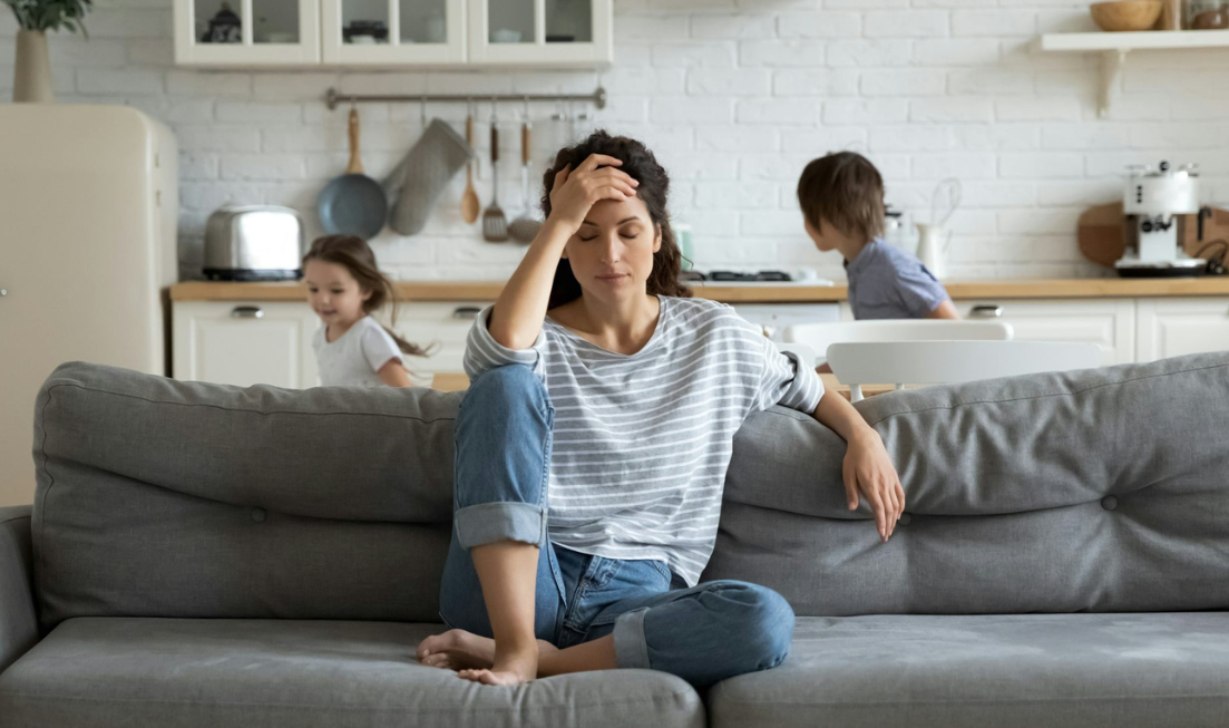 A woman holds her hand to her head. She looks stressed as she sits in a living room.