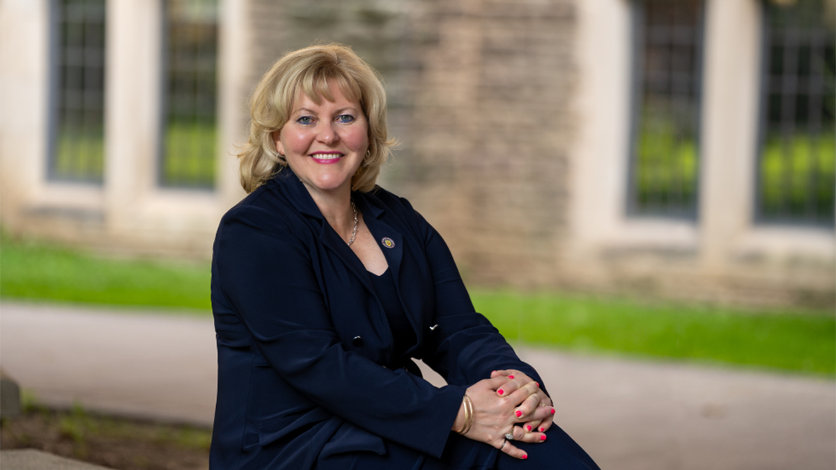 A woman in a navy suit with blond hair, faces camera with a smile. She is sitting on a bench with stonework and green grass behind her.