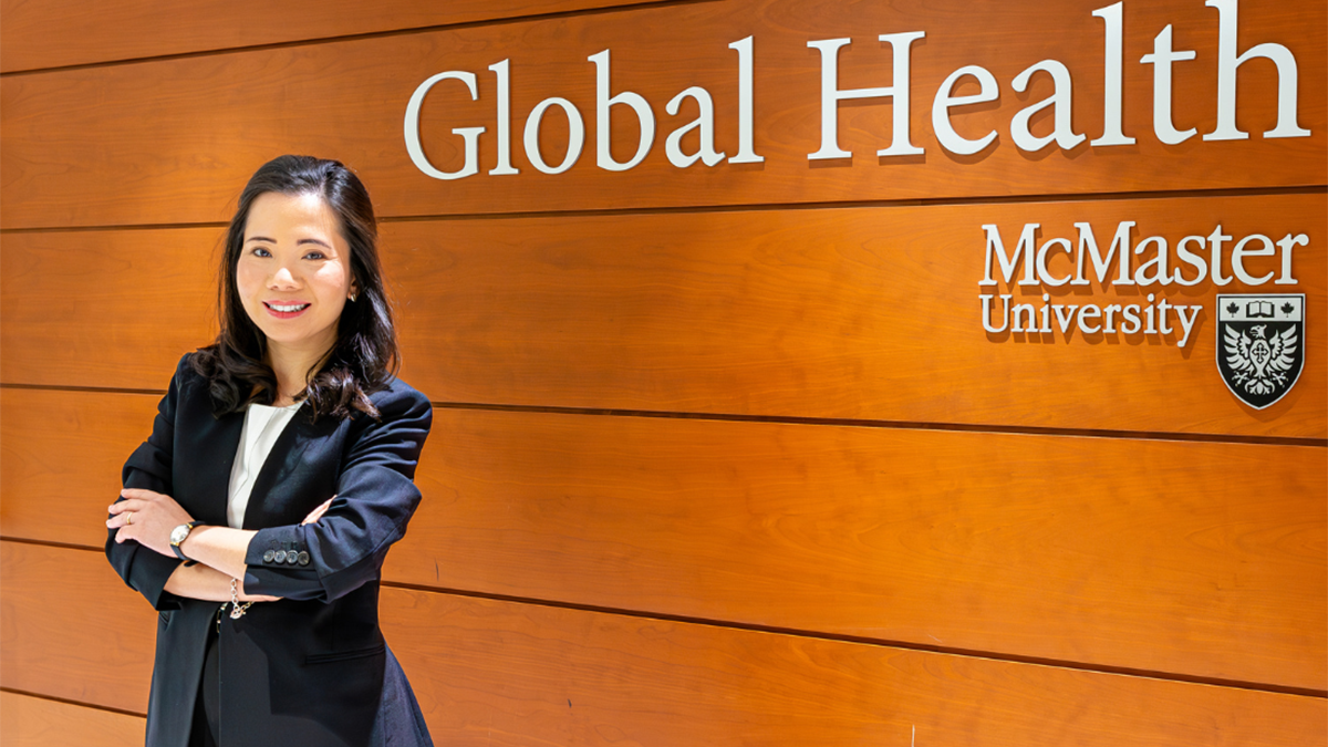 A smiling person in a suit standing with their arms crossed in front of the McMaster Global Health sign in the Faculty of Health Sciences.