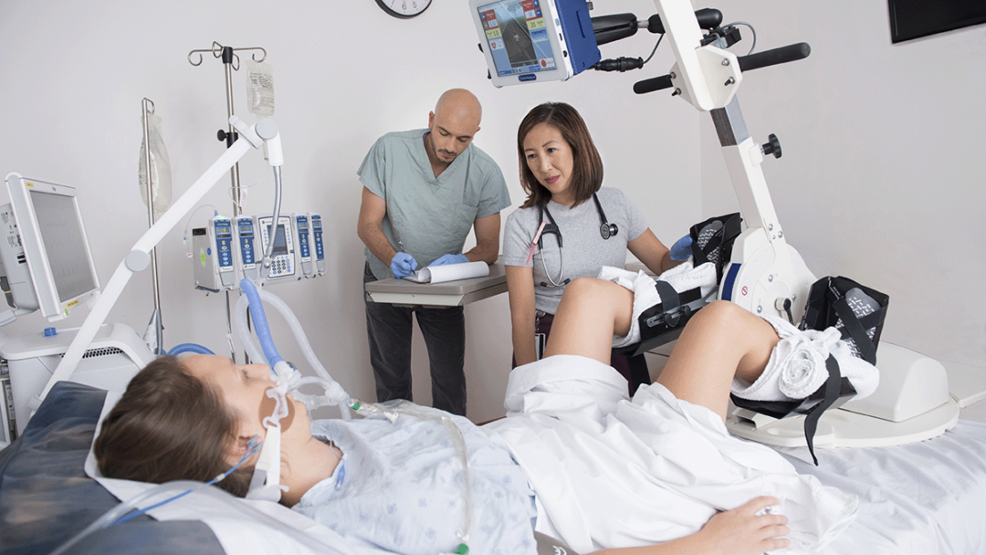 Researcher Michelle Kho stands at the end of a bed where a person, laying in a bed, is simulating the use of a cycling machine for the purpose of rehab.