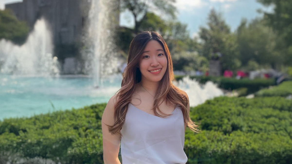 Ashley Jong can be seen standing in front of a water fountain, with lush greenery surrounding it. Jong is wearing a grey shirt and beige pants.