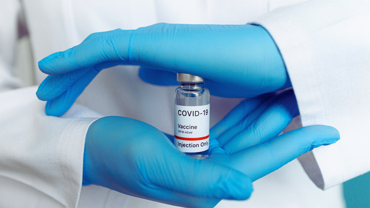 A person wearing blue gloves holds a vile containing with a label identifying it as a COVID-19 vaccine.