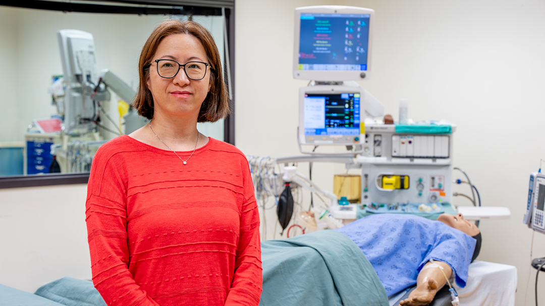 Bingxian Wang stands in a bright red shirt in the Centre for Simulation Based Learning. Behind her is a full-body patient simulator – also called a manikin – on a hospital bed with medical equipment.