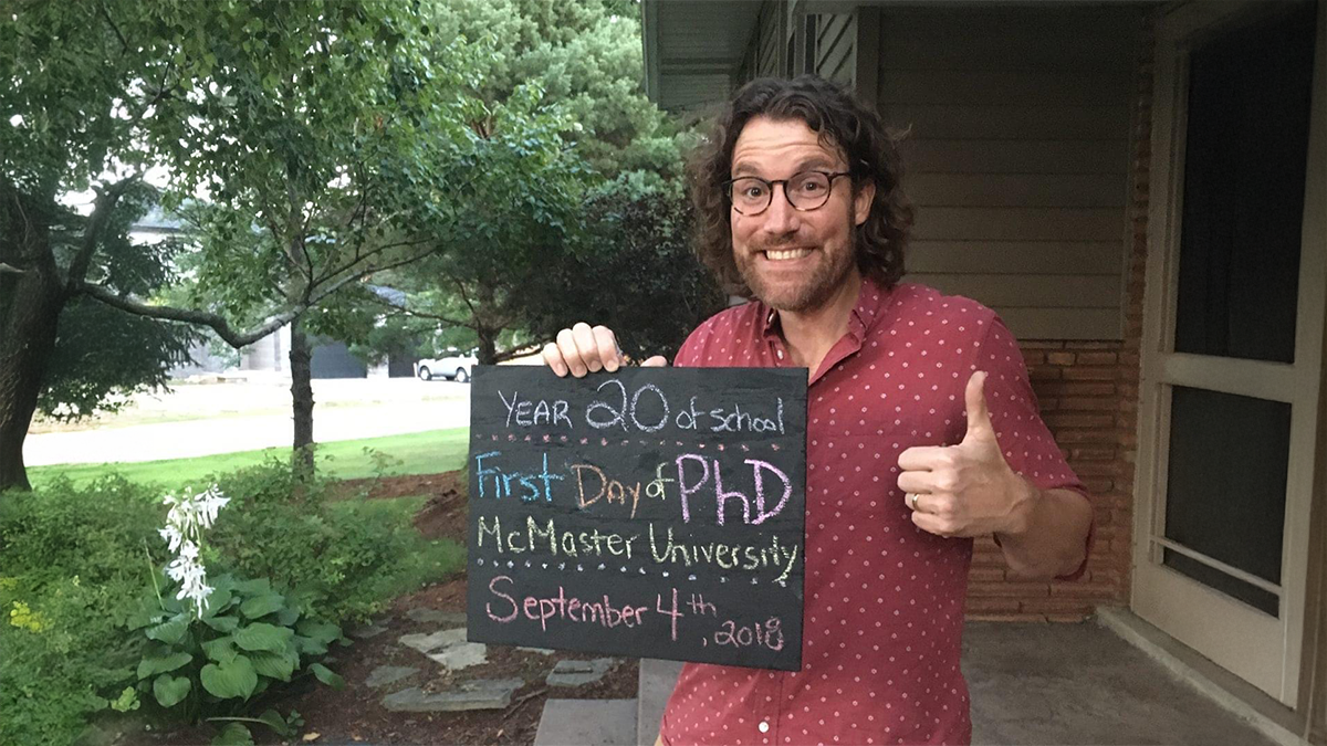 Aric Rankin holds a chalk board sign about the first day of his PhD, like a child would on their first day of school.