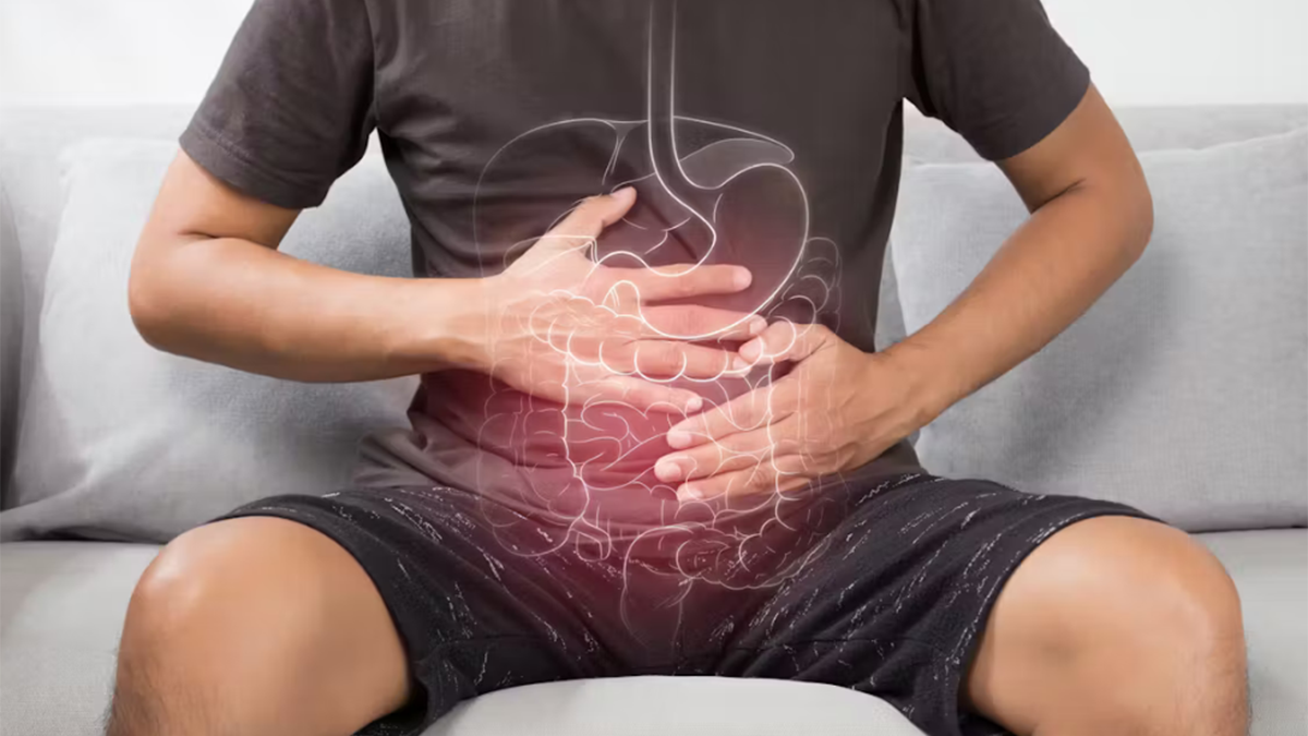 A photo showing the midsection of person sitting on a grey couch in a black t-shirt and shorts. Their hands are resting on their abdomen and there is a graphic design of internal organs overlaid over the person’s stomach area.