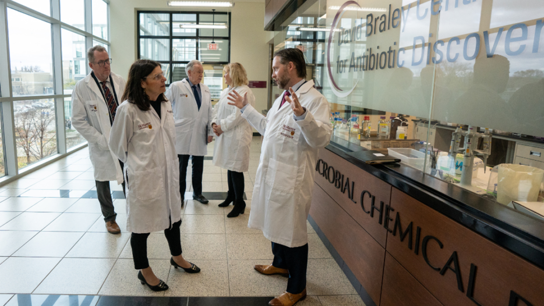 University and government personnel speak together in a corridor outside a McMaster laboratory. The five people pictured are all wearing lab coats.