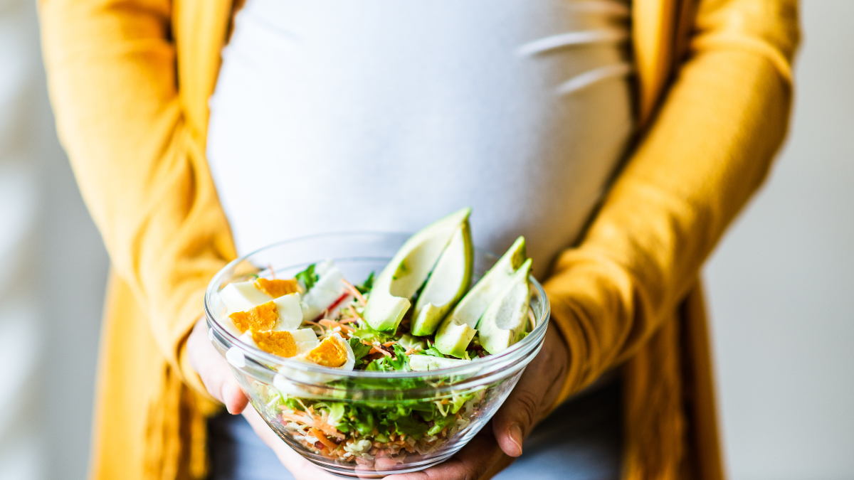 A pregnant person wearing a white shirt and a yellow sweater is holding a bowl of healthy food, including leafy greens, boiled eggs and avocados.