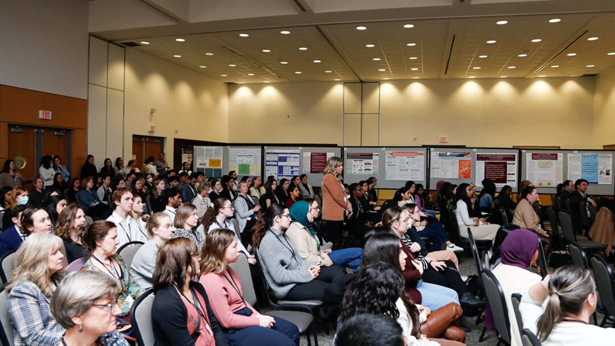 A large audience in a well-lit room attentively views research posters on the walls.