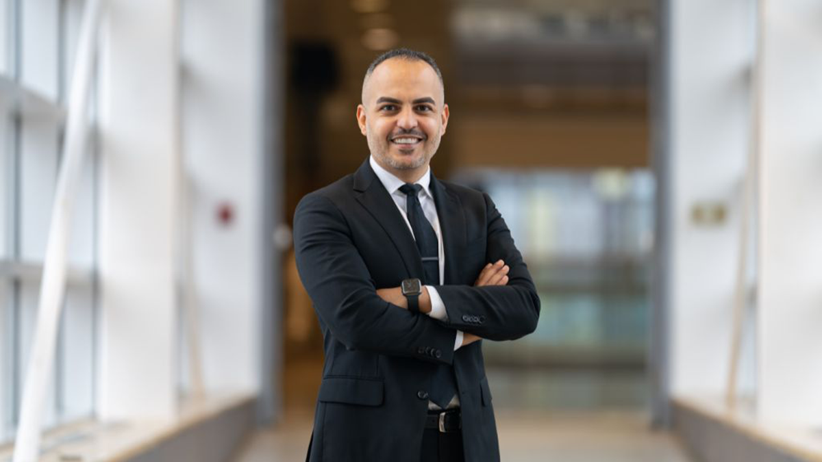 Firas Khalid smiles confidently indoors, wearing a white shirt, black blazer, and tie.