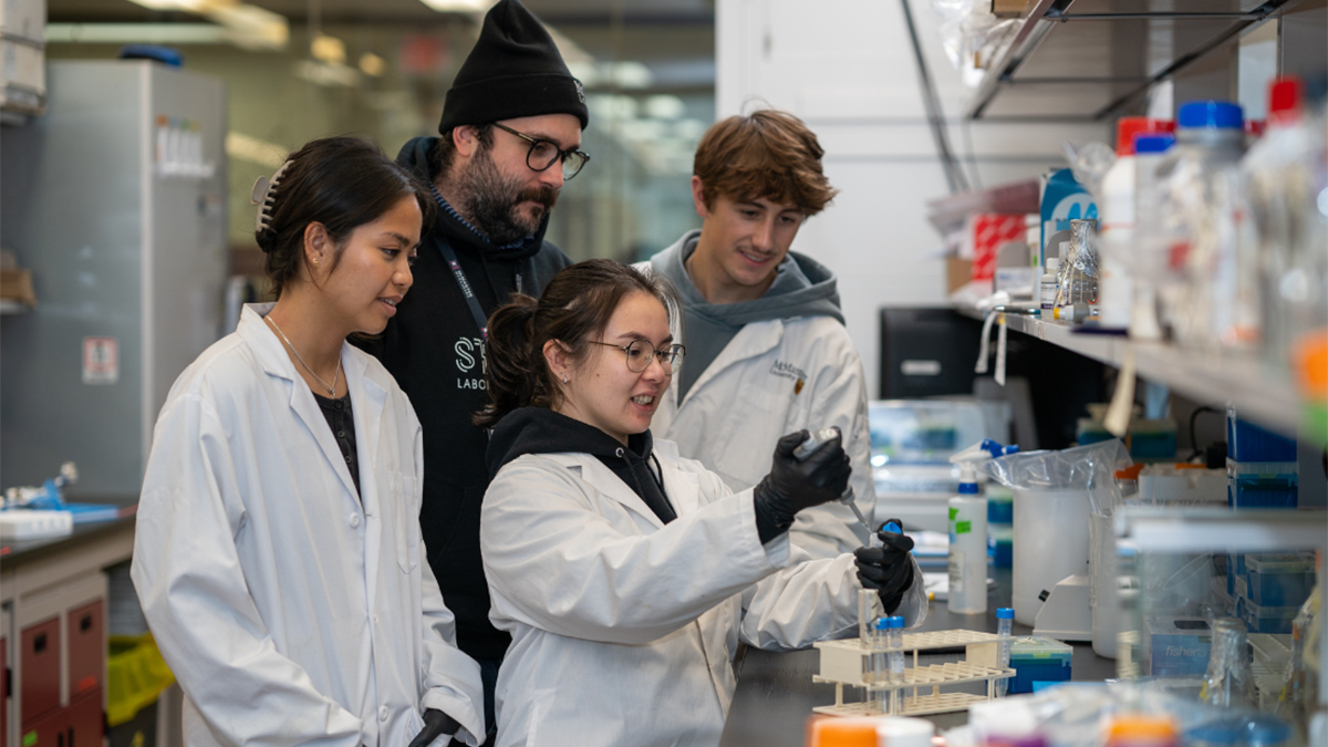 A grad student works with a pipette and beaker in a McMaster lab, observed by a professor and two others.