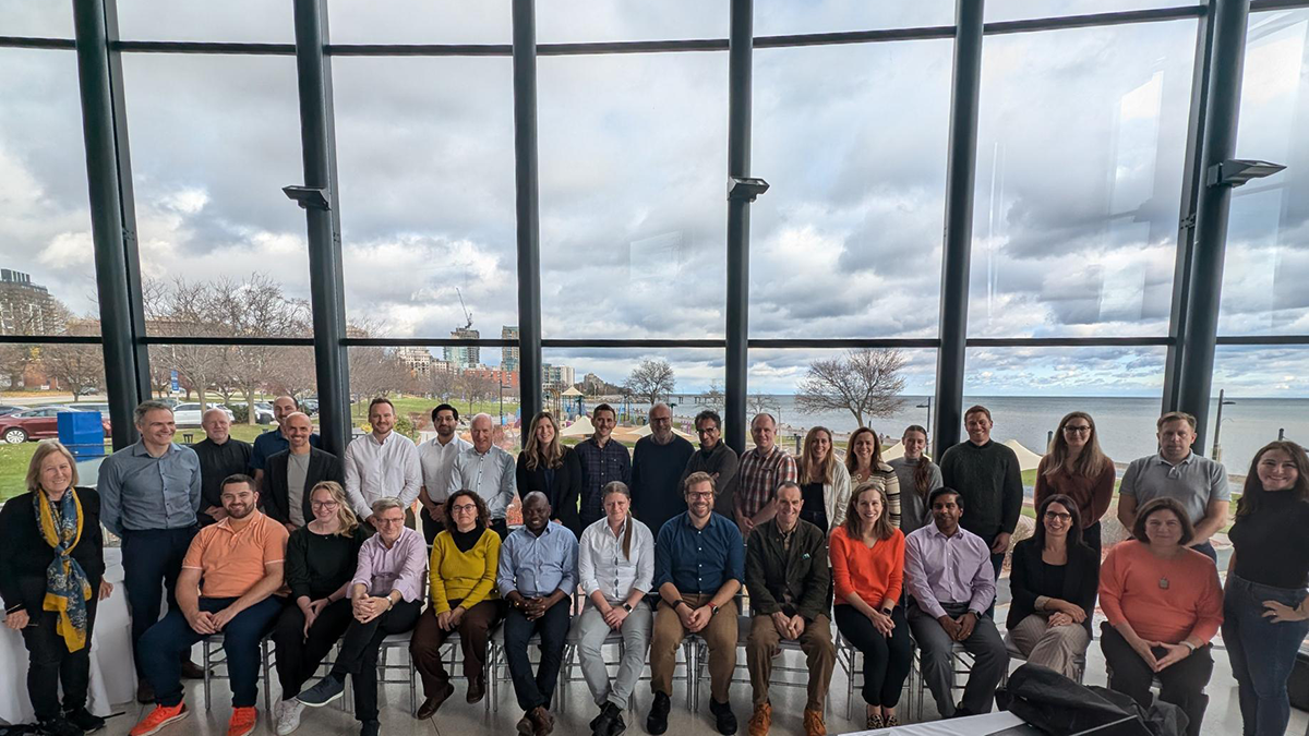 A diverse group poses in front of windows with a waterfront and cloudy sky in the background.