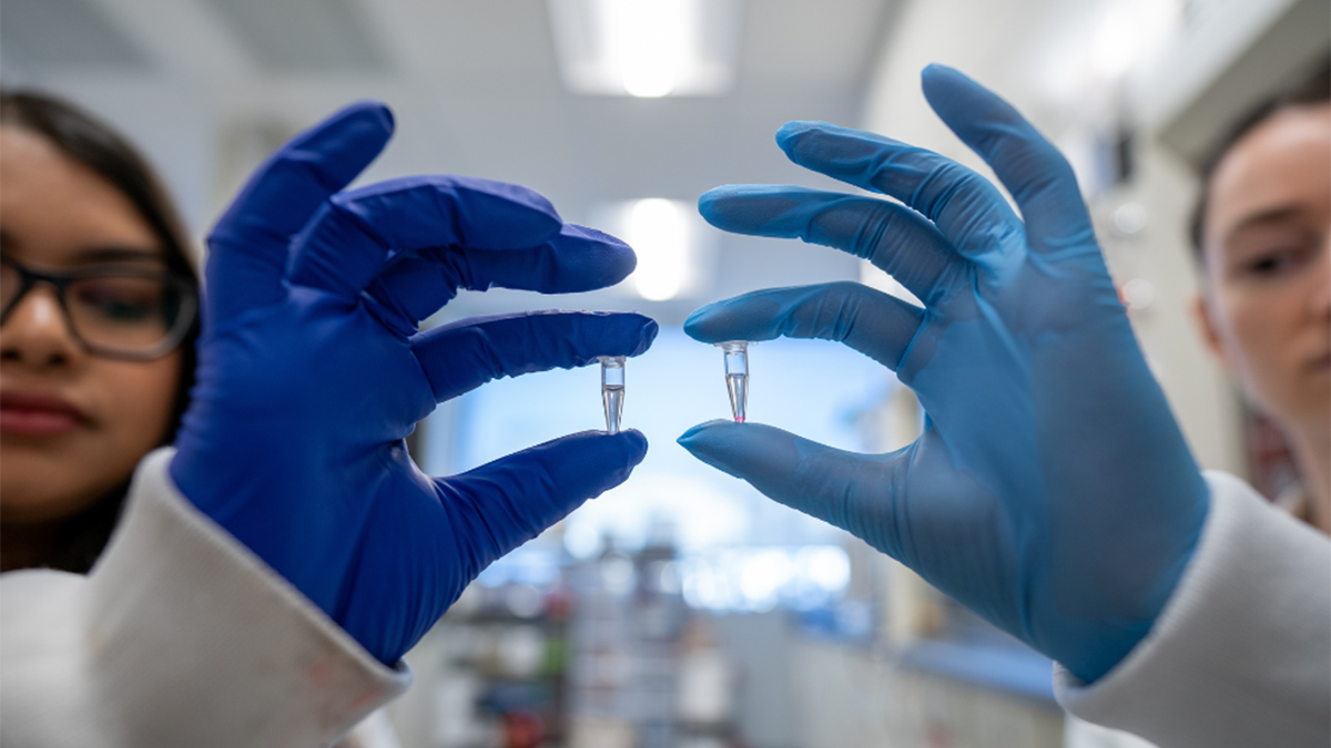 Two gloved hands holding small vials of liquid, with two blurred faces in the background.