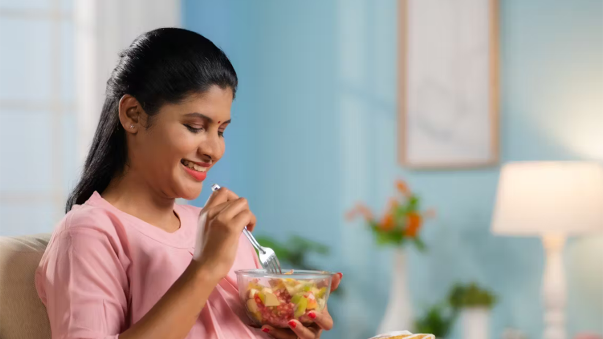 Woman in pink shirt eating fruit from a glass bowl with a fork.