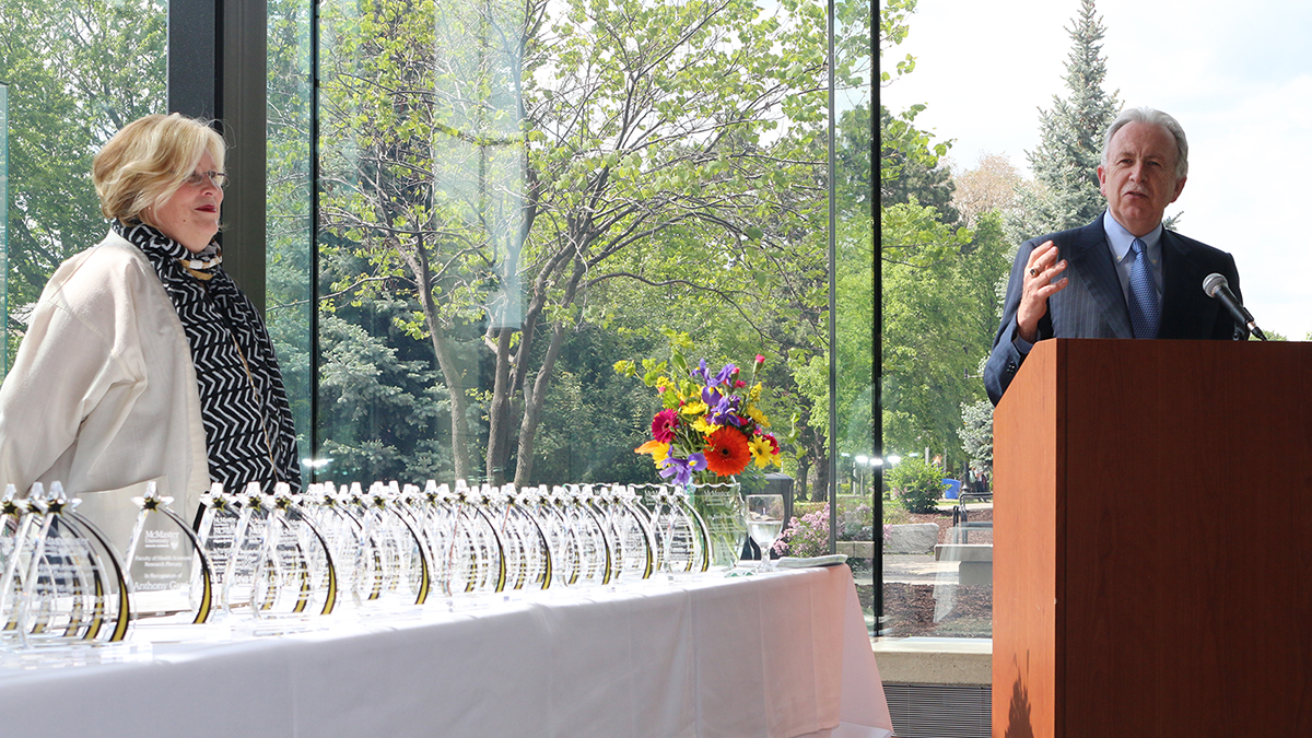 Catherine Hayward is pictured on stage behind a row of personalized glass award plaques. To her right is Paul O’Byrne, pictured at a podium.