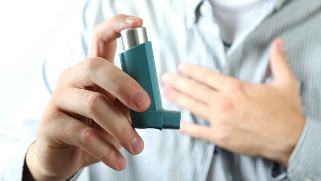 A person holds an inhaler with one hand and has their other hand over their chest.