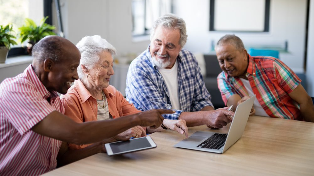 Four older adults, in discussion, sitting at table with laptop and tablet.