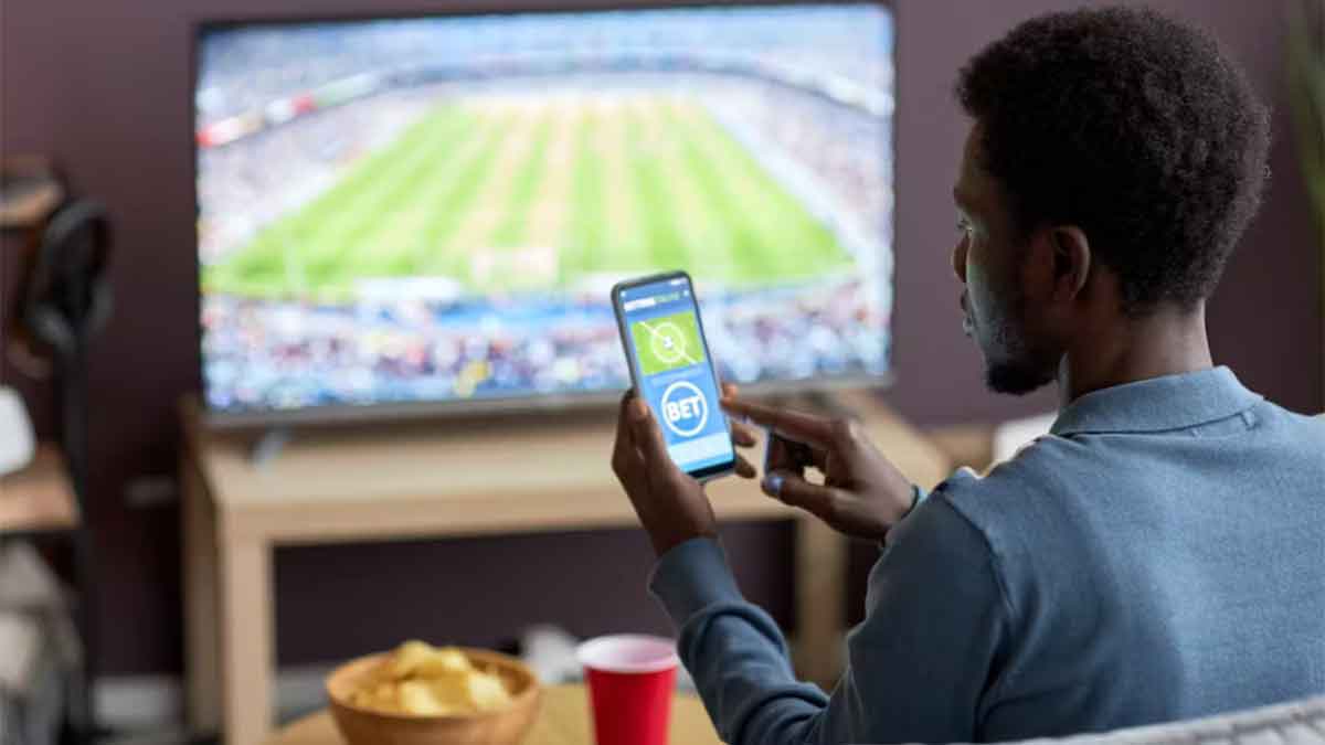 Person looking at a cellphone with a TV in the background showing a sporting event.