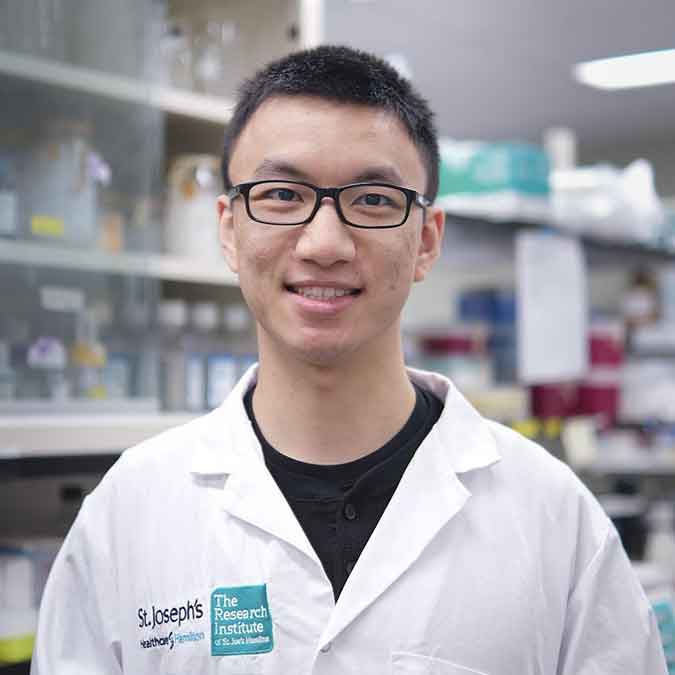 Tim Ju smiles in laboratory wearing a white lab coat.